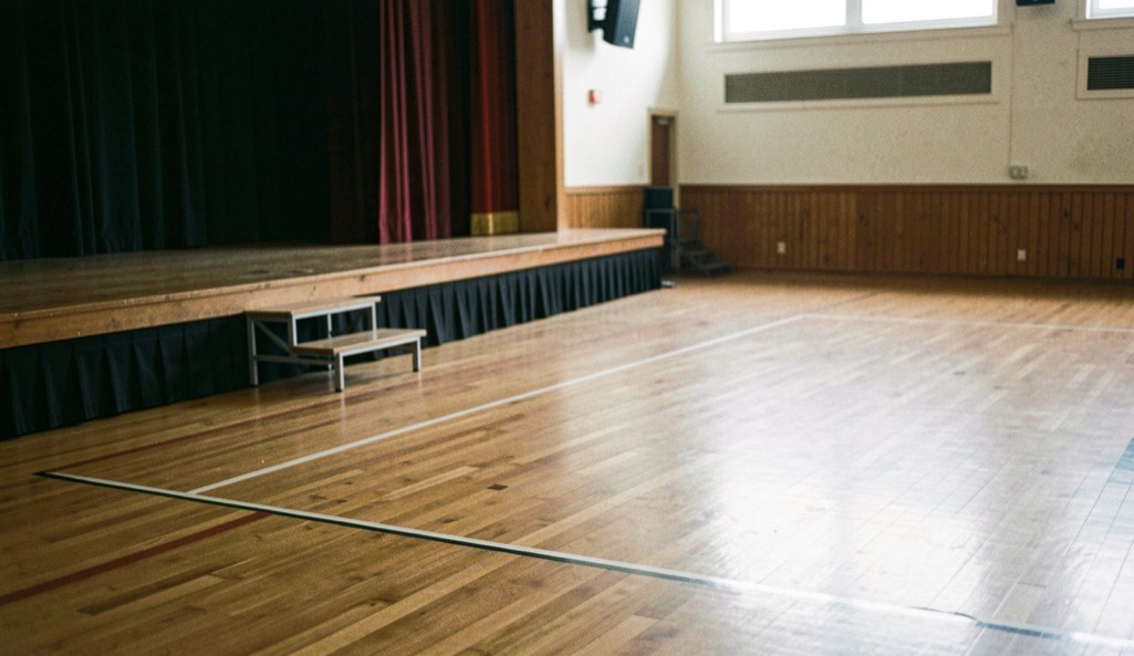 An empty hardwood ballroom floor with a small stage and clear walkways.